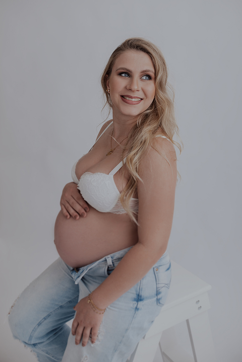 Foto de uma gestante em ensaio fotográfico, sorrindo e apoiando uma das mãos sobre a barriga.